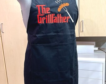Könnte beinhalten: Schwarze Schürze mit einem großen roten Stickmuster, auf dem "The Grillfather" steht, darüber ein Cartoon-Bild eines Hotdogs auf einer Gabel.
