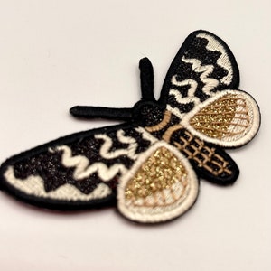 May include: A black and white embroidered moth with gold glitter accents. The moth has a detailed design with a white outline and a black body. The wings are spread out and the moth is facing to the right.