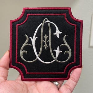 May include: Embroidered patch with a black background and a red border. The patch features the intertwined letters "A" and "C" in white, with gray and white decorative flourishes. The patch is square with rounded corners.