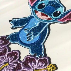 Op de afbeelding: Blauw en paars glitter strijk-op patch met Stitch van Lilo & Stitch die op een paarse hibiscus bloem staat.