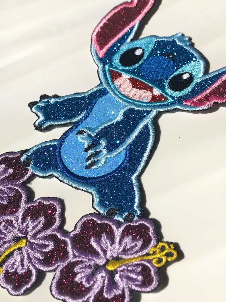 Op de afbeelding: Blauw en paars glitter strijk-op patch met Stitch van Lilo & Stitch die op een paarse hibiscus bloem staat.