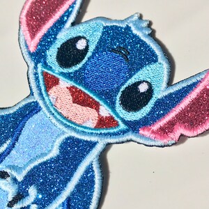 Op de afbeelding: Een blauwe en roze glitter strijk-op patch met Stitch uit de Disney film Lilo & Stitch. Stitch glimlacht met zijn kenmerkende ondeugende uitdrukking.