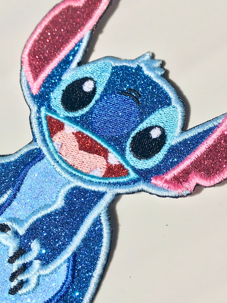 Op de afbeelding: Een blauwe en roze glitter strijk-op patch met Stitch uit de Disney film Lilo & Stitch. Stitch glimlacht met zijn kenmerkende ondeugende uitdrukking.