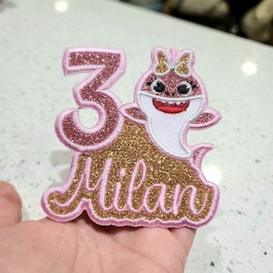 May include: Pink and gold glitter iron-on patch with a cartoon baby shark wearing a bow, the number 3, and the name Milan.