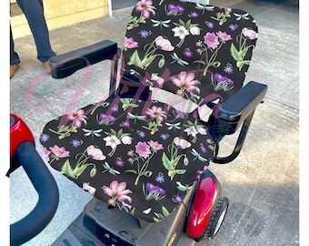 Puede incluir: Funda de asiento de scooter de movilidad negra con un patrón floral que presenta flores rosas, blancas y moradas, hojas verdes y detalles de libélulas.