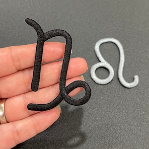 Op de afbeelding: Twee sterrenbeeld patches, een zwarte en een zilveren. De zwarte patch is een Boogschutter symbool en de zilveren patch is een Leeuw symbool.