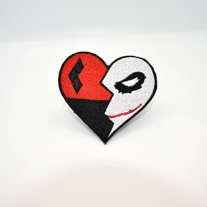 May include: A red and black heart-shaped patch with a diamond pattern on one side and a white Joker face on the other side.