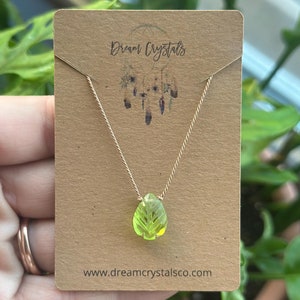 May include: A delicate gold chain necklace with a green pear-shaped pendant. The pendant has a carved leaf design. The necklace is displayed on a brown card with the text "Dream Crystals" and "www.dreamcrystalsco.com".