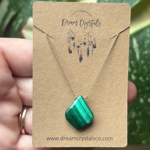 May include: A green malachite gemstone pendant necklace. The gemstone is diamond-shaped and hangs from a delicate gold-colored chain. The necklace is displayed on a brown card with the text "Dream Crystals" and "www.dreamcrystalsco.com".