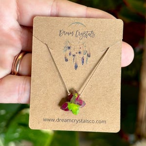 May include: A necklace featuring a small, butterfly-shaped pendant with vibrant magenta and lime green coloring, suspended on a thin tan cord. The necklace is displayed on a rectangular brown card printed with the text Dream Crystals.