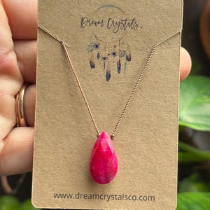 May include: A faceted, teardrop-shaped hot pink gemstone pendant on a thin, twisted metallic cord. The necklace is displayed on a rectangular tan card featuring the text Dream Crystals and the website www.dreamcrystalsco.com.
