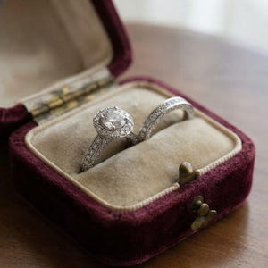 May include: A silver-toned engagement ring featuring a round center stone surrounded by a halo of smaller stones, paired with a matching curved wedding band. Both rings are set with pavé stones and displayed inside a deep burgundy velvet box.