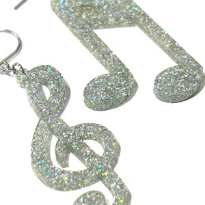 May include: A pair of silver glitter earrings, one shaped like a treble clef and the other a musical note. The earrings have a sparkly, iridescent finish and are suspended from silver ear wires. The musical-themed jewellery is set against a white background.