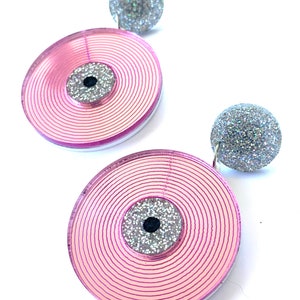 Op de afbeelding: Een paar roze en zilveren oorbellen. Elke oorbel heeft een grote, cirkelvormige roze schijf met concentrische lijnen, een zilveren glittercentrum en een kleinere zilveren glitterbal aan de bovenkant. De oorbellen zijn voor doorboorde oren.