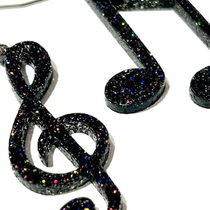 May include: A pair of black glitter earrings in the shape of a music note and a treble clef. The earrings are made of a dark material with colourful glitter. The treble clef is on the left, and the music note is on the right.