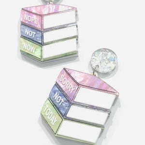 Peut inclure: Boucles d'oreilles fantaisie composées d'une pile de trois livres avec les phrases "Nope, Not, Now" et "Sorry, Not, Today" en texte blanc. Les boucles d'oreilles présentent un dégradé de rose, bleu et vert nacré, surmontées d'une gemme transparente à facettes.