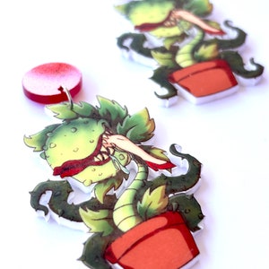 May include: Novelty earrings with a cartoon design of a carnivorous plant. The earrings show a green plant with a red mouth and a red pot. The earrings are made of a clear material with a red circular top.