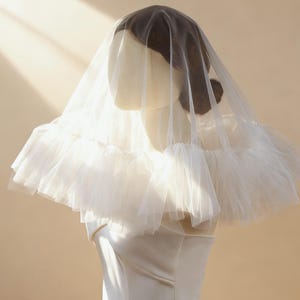 Puede incluir: Un velo de novia blanco con un diseño en capas y con volantes. El velo está hecho de una tela fina y delicada y está drapeado sobre una cabeza de maniquí. El maniquí lleva un vestido blanco. El fondo es de un color suave y neutro.