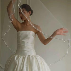 May include: A white strapless wedding dress with a full skirt. The bride is holding a sheer veil edged with pearls. The veil is draped over her head and arms. The dress is made of a satin-like material.