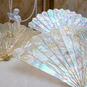 Può includere: Due ventagli pieghevoli in madreperla iridescente. I ventagli sono chiusi e hanno un design delicato e decorato.