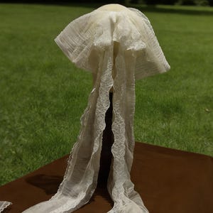 May include: An ivory vintage veil with a wide, pleated brim and long, flowing lace trim. The veil is displayed on a dark brown surface against a backdrop of green grass and trees.
