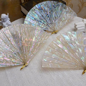 May include: Three iridescent mother-of-pearl folding fans with gold accents. The fans are closed and laid out on a white surface.