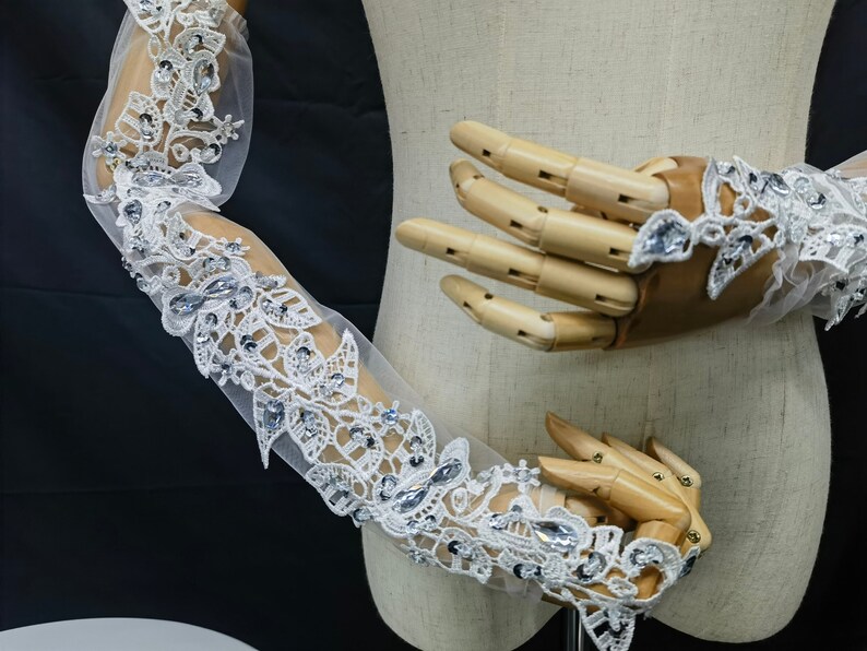 May include: Long white lace gloves with floral patterns and sparkling rhinestones. The gloves extend up the arm and are displayed on a wooden mannequin hand. The design features delicate lace and shimmering embellishments.