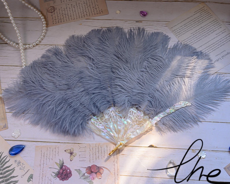 K&ouml;nnte beinhalten: Hellblauer Federf&auml;cher mit dekorativem Griff. Der F&auml;cher besteht aus weichen, flauschigen Federn und hat einen perlmuttartigen Griff. Der F&auml;cher liegt auf einer Holzoberfl&auml;che mit Vintage-Papier und dekorativen Elementen.