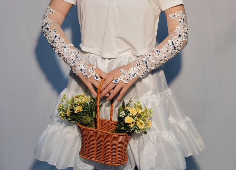 May include: Long white lace gloves with blue embellishments and floral patterns. A brown wicker basket, filled with yellow and green flowers, is held in front of a white dress. The gloves cover the arms.