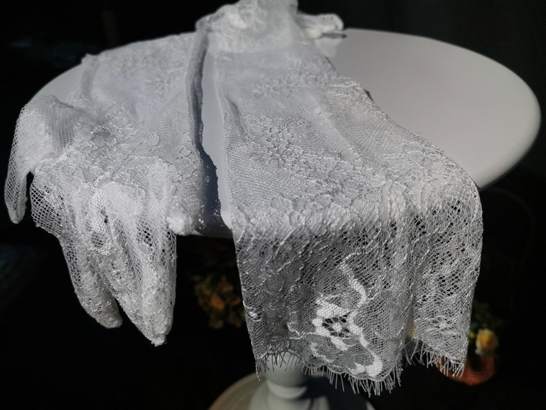 May include: A pair of white lace gloves with a delicate floral pattern. The gloves are made of a soft, sheer fabric and have a scalloped edge.