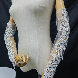 May include: Elegant sheer white gloves for a bride, adorned with detailed floral lace and shimmering crystal accents. The long gloves are displayed on a mannequin. The design showcases delicate patterns and a touch of glamour for a special occasion.