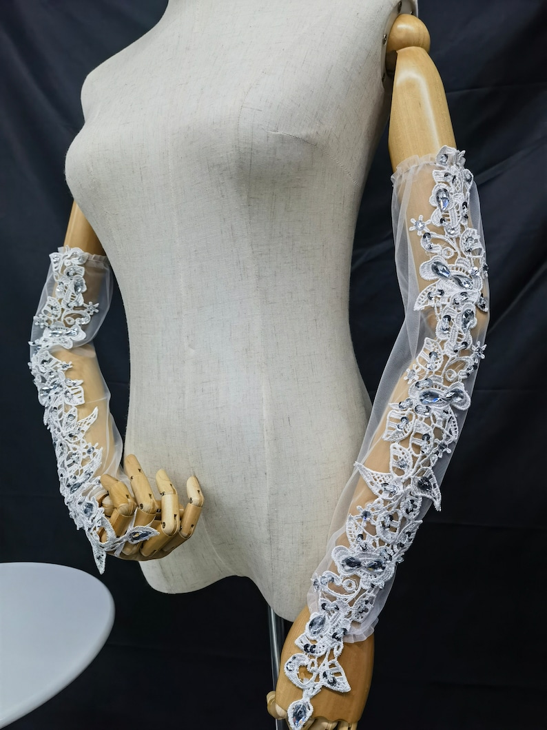 May include: Elegant sheer white gloves for a bride, adorned with detailed floral lace and shimmering crystal accents. The long gloves are displayed on a mannequin. The design showcases delicate patterns and a touch of glamour for a special occasion.