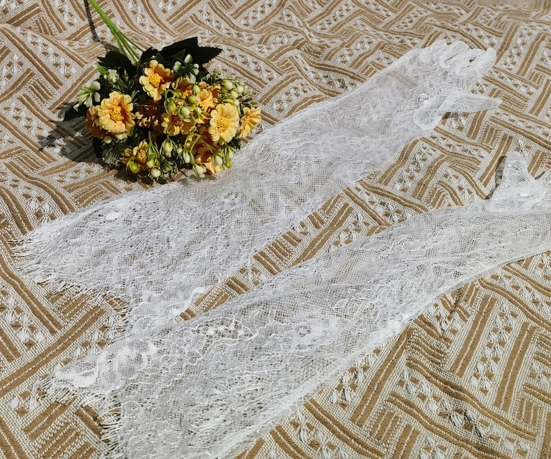 May include: Two white lace gloves on a brown patterned fabric background. The gloves are made of delicate lace and have a soft, feminine look.