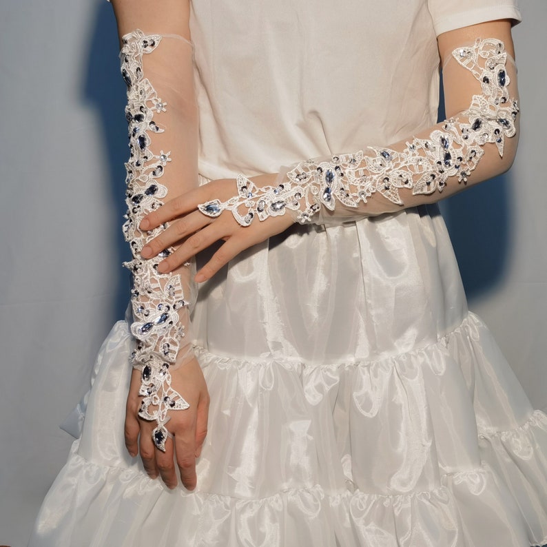May include: Elegant sheer white gloves with delicate floral lace and sparkling blue gemstones. The gloves reach the upper arm and include a finger ring, offering a sophisticated appearance. Ideal for weddings or special events.