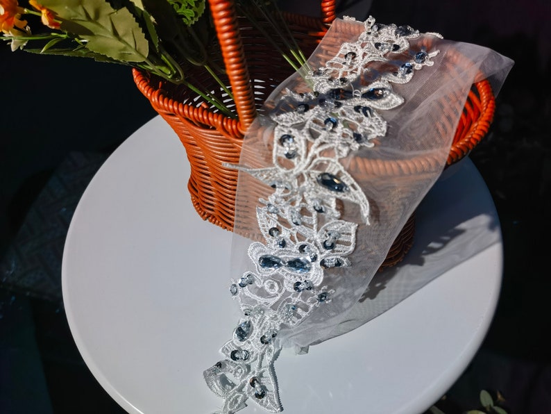 May include: A white lace trim with a floral pattern, adorned with dark blue gemstones. The lace is set on a sheer, white fabric, creating a layered effect. The trim is suitable for bridal or formal attire.