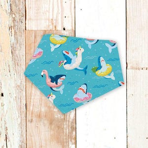 May include: A turquoise bandana with a playful pattern of sharks, flamingos, and unicorns in inflatable rings. The design includes wavy lines, suggesting water, and is set against a light wood background.