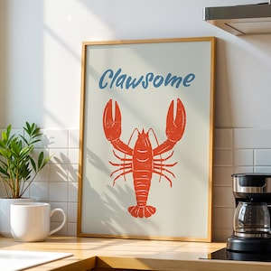May include: A framed art print featuring a red lobster illustration with the word "Clawsome" in blue script. The artwork is displayed on a kitchen counter, alongside a coffee maker and a potted plant.