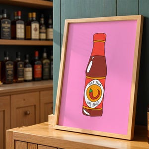 Op de afbeelding: Een ingelijste print met een cartoon illustratie van een hete sausfles op een roze achtergrond. De fles is rood en bruin met een label met de tekst "hey there hot stuff" en een chilipeperafbeelding. De lijst is licht van kleur.