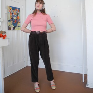 May include: A woman wearing a light pink short-sleeved top with small pearl-like buttons, paired with dark brown corduroy pants and striped sandals. A patterned scarf is tied around the head. The outfit is completed with a brown belt.