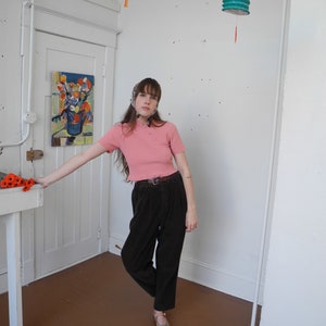 May include: A person wearing a pink short-sleeved top with small pearl-like buttons, dark brown pleated pants, and striped sandals. The person is standing in a room with white walls and a painting on the wall. Two paper lanterns hang from the ceiling.