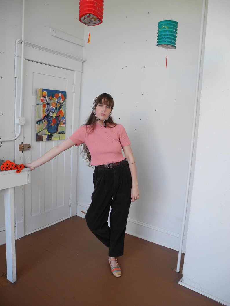 May include: A person wearing a pink short-sleeved top with small pearl-like buttons, dark brown pleated pants, and striped sandals. The person is standing in a room with white walls and a painting on the wall. Two paper lanterns hang from the ceiling.