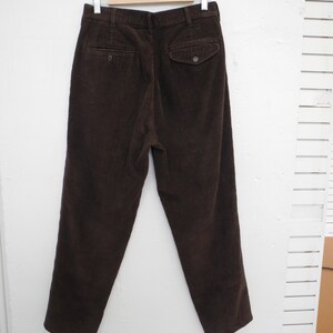 May include: Dark brown corduroy pants hanging on a wooden hanger. The pants have two back pockets with button closures and a classic, tailored fit. The corduroy fabric has a ribbed texture.