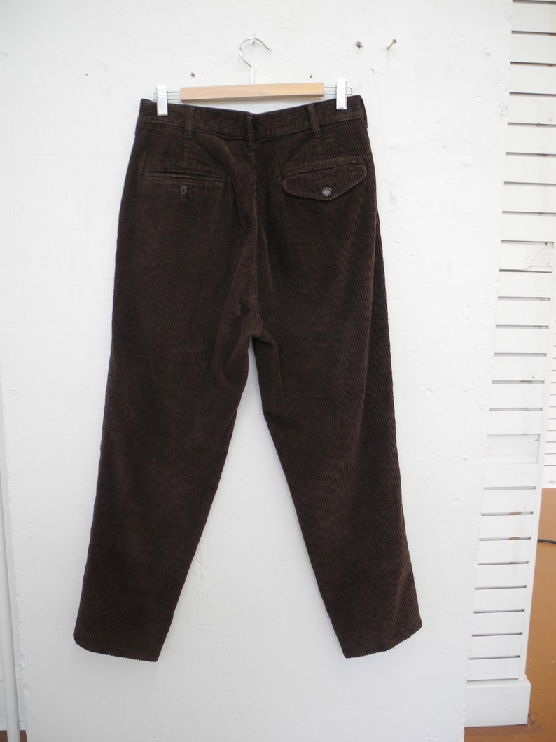 May include: Dark brown corduroy pants hanging on a wooden hanger. The pants have two back pockets with button closures and a classic, tailored fit. The corduroy fabric has a ribbed texture.