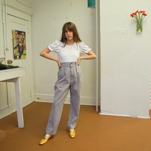 May include: A woman wearing a white top with puffed sleeves and high-waisted striped pants. She is standing in a room with white walls and a wooden floor. There are three paper lanterns hanging from the ceiling.