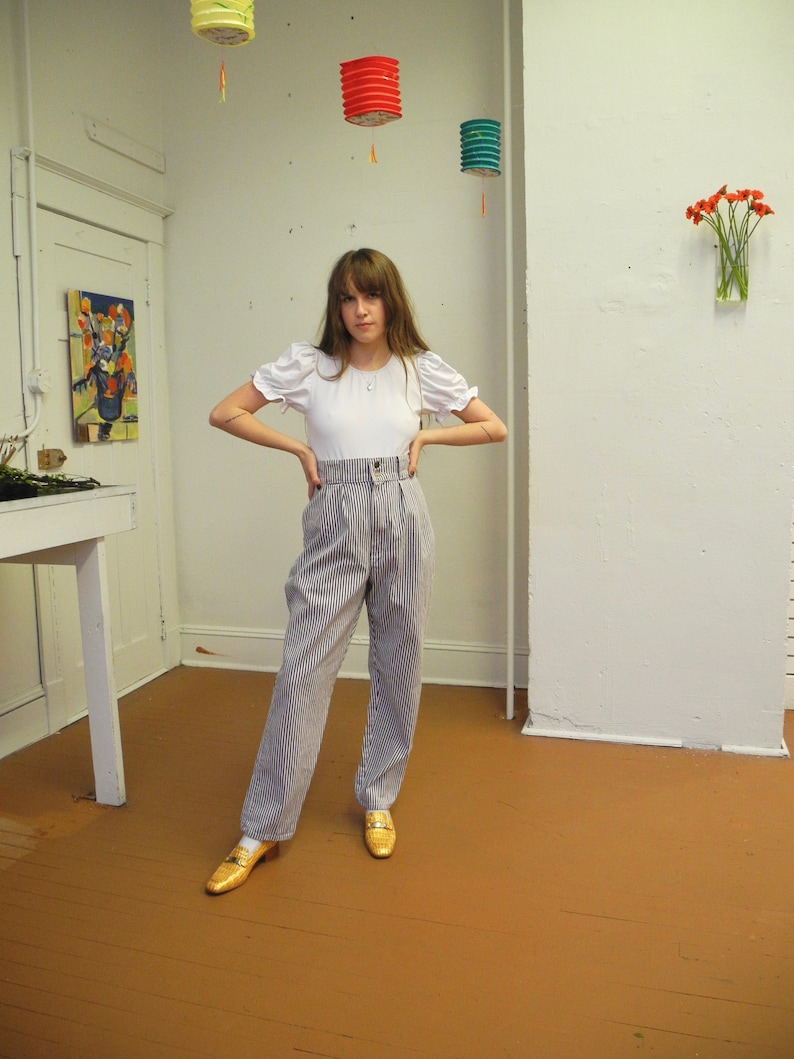 May include: A woman wearing a white top with puffed sleeves and high-waisted striped pants. She is standing in a room with white walls and a wooden floor. There are three paper lanterns hanging from the ceiling.