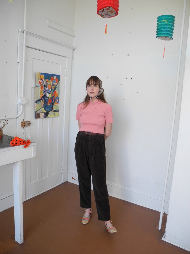 May include: A woman wearing a pink short-sleeved shirt, dark brown corduroy pants, and striped sandals. She is standing in a room with white walls, a white door, and a painting on the wall. Decorative paper lanterns hang from the ceiling.