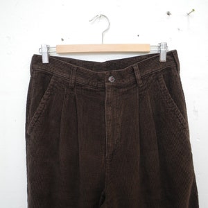 May include: Close-up of brown corduroy pants hanging on a wooden hanger. The pants have a button closure, pockets, and pleats at the waist. The corduroy fabric has a ribbed texture.