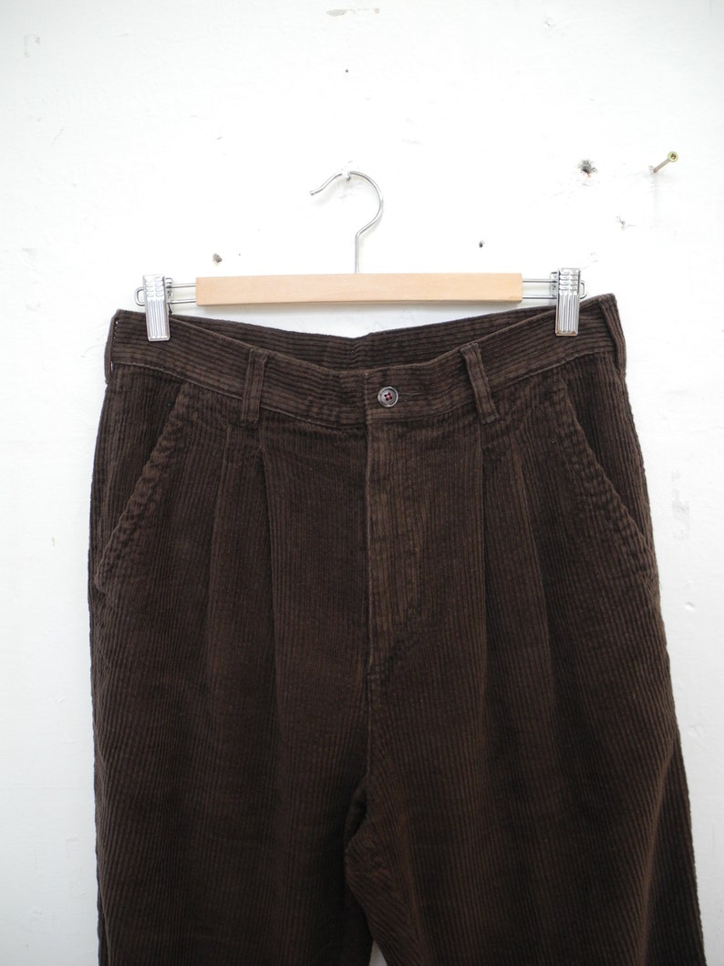 May include: Close-up of brown corduroy pants hanging on a wooden hanger. The pants have a button closure, pockets, and pleats at the waist. The corduroy fabric has a ribbed texture.