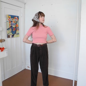 May include: A person wearing a pink short-sleeved top with small pearl-like buttons, brown corduroy pants, and striped sandals. A patterned scarf is tied around the head. The outfit is completed with a brown belt.