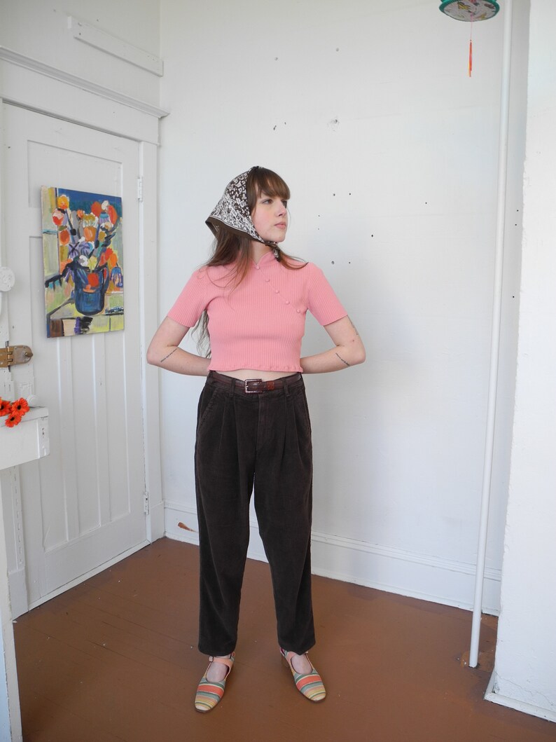 May include: A person wearing a pink short-sleeved top with small pearl-like buttons, brown corduroy pants, and striped sandals. A patterned scarf is tied around the head. The outfit is completed with a brown belt.